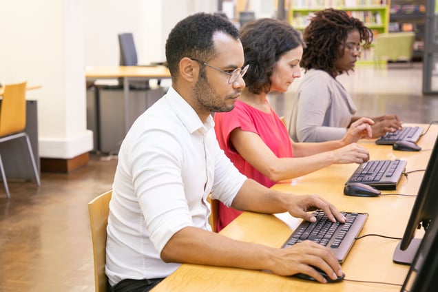 Three employees sitting at the same table, working on their computers. The employee on the left has glasses, a white shirt, and a low beard. The employee in the middle is a woman with black hair, wearing a pink T-shirt. The employee on the right has glasses and is wearing a grey sweater.