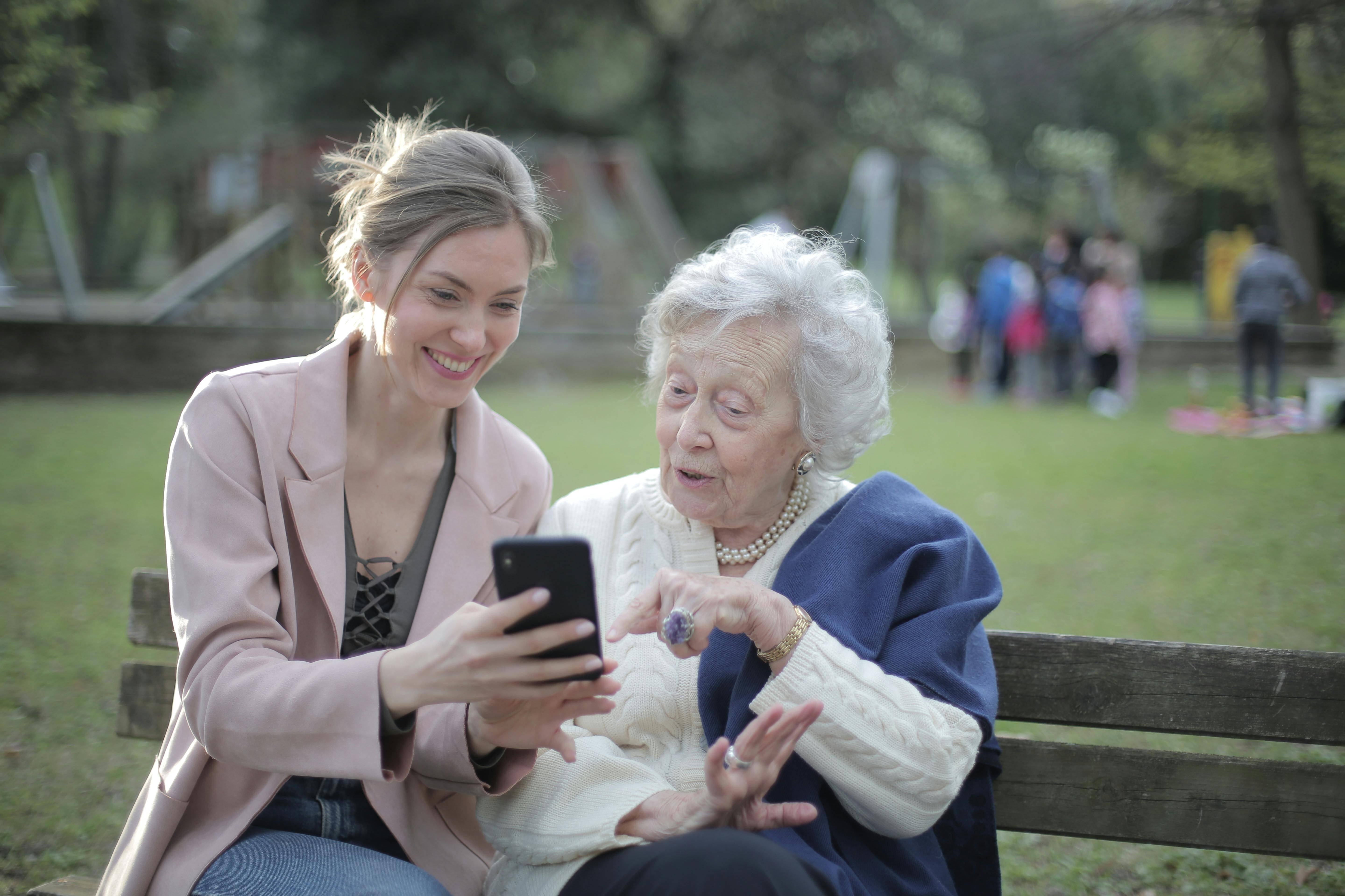 A younger white woman (left) is sat on a bench with an elderly white man (right) showing her something on a mobile phone.