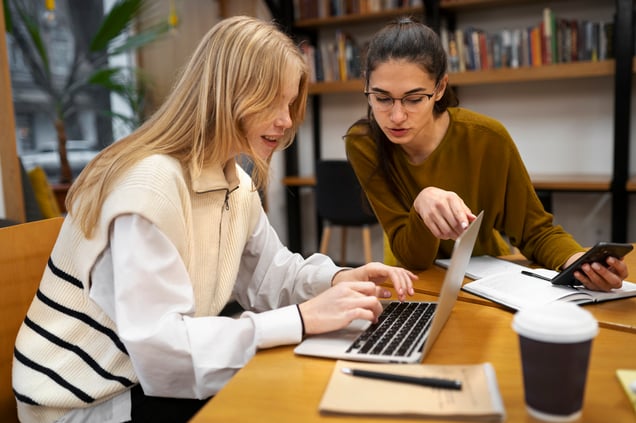 Two women sitting at a table, working together on a laptop. The woman on the right is holding her phone while they both focus on the screen.